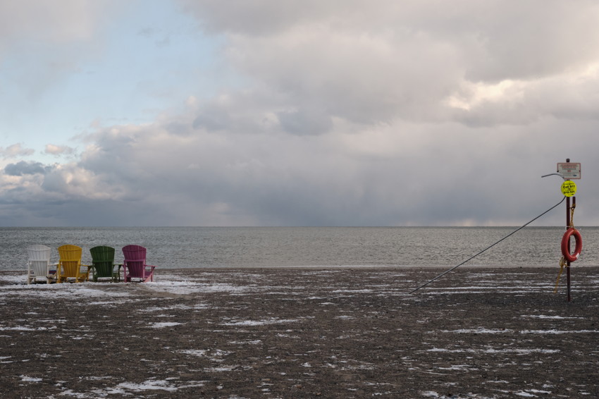Four empty colorful Muskoka chairs on snowy beach, flat lake horizon, lifebuoy on pole at right.