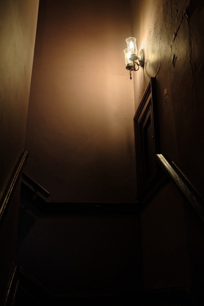 Dim narrow stairwell viewed upward, wall sconce with two bulbs casting light, peeling plaster, wooden banister.