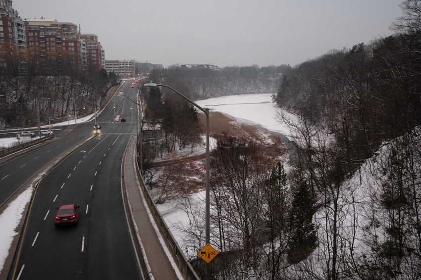 Elevated view of multi-lane road with cars left, streetlight center, snowy wooded ravine and frozen pond right.