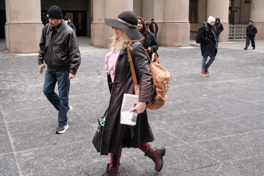 Person walking in city plaza wearing wide-brim hat, long coat, plaid leggings, tan backpack, holding folded newspaper.