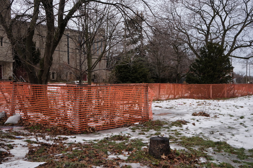Orange construction fence around trees and patchy snow, tree stump in foreground, building behind.