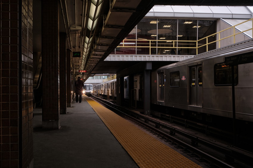 Train platform with yellow tactile strip, person waiting, approaching train headlight, stationary train on right.