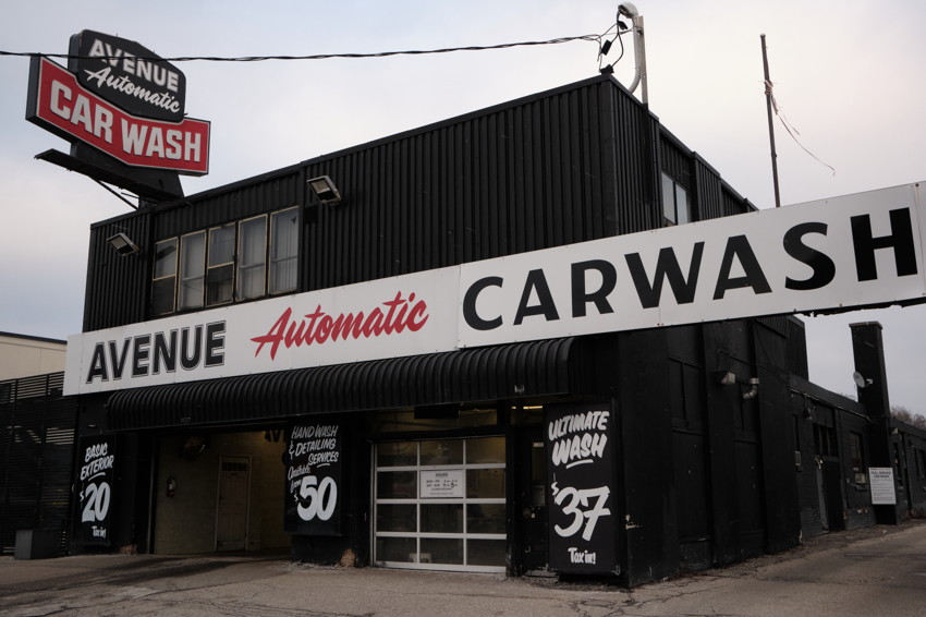 Corner view of black car wash building with large white AVENUE Automatic CARWASH sign and visible price boards.