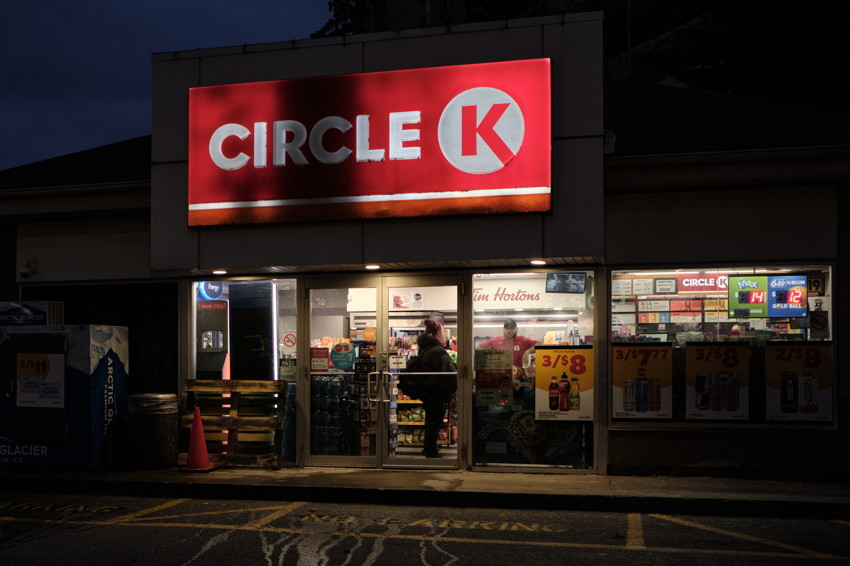 Frontal nighttime view of illuminated Circle K storefront with glass doors, person inside, and promotional signage.