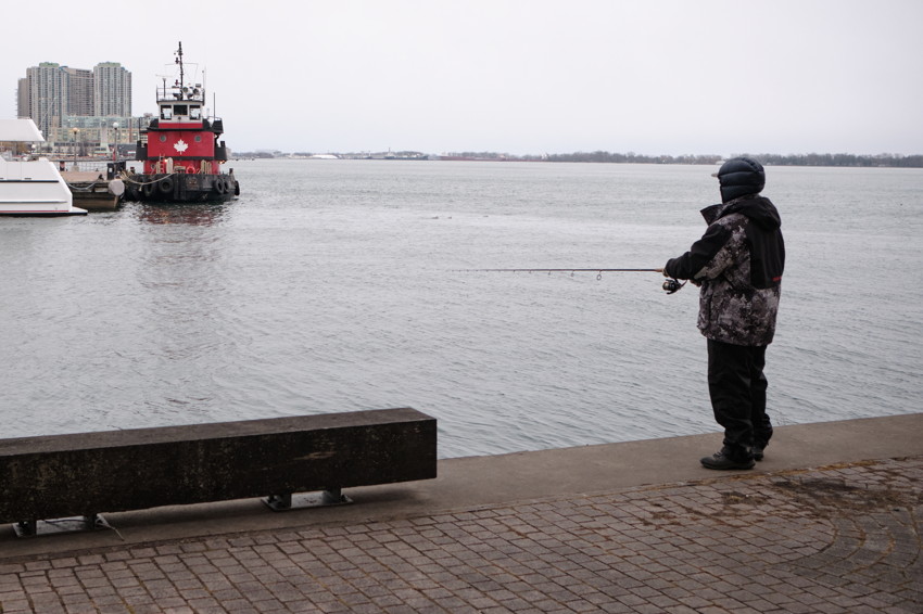 Person in winter coat fishing from quay, rod extended; red tugboat and skyline left, bench foreground.