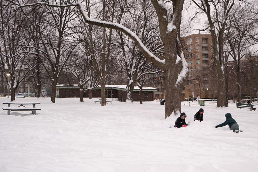 Snow-covered urban park with large bare trees, picnic tables left, three children playing in deep snow right.