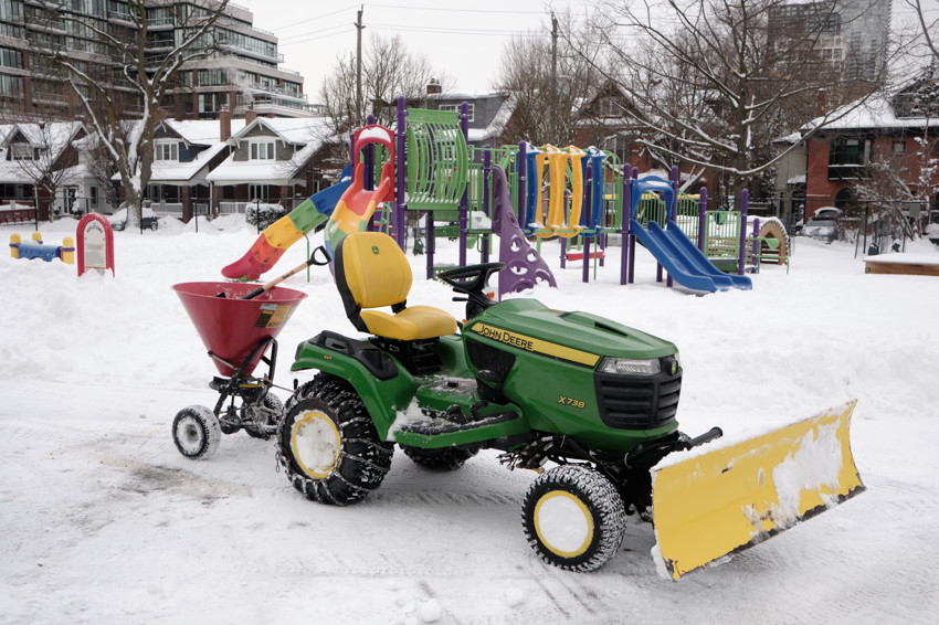 Green John Deere tractor with yellow snowplow and red spreader in snowy playground with colorful play structure.