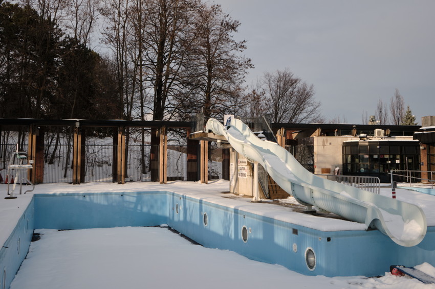 Empty outdoor pool with snow, blue tiled basin and white curving waterslide under leafless trees.