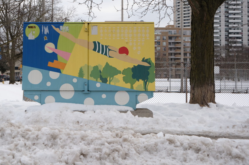 Painted utility box showing a diver and abstract trees; snow-covered ground, tree trunk and apartment buildings behind.