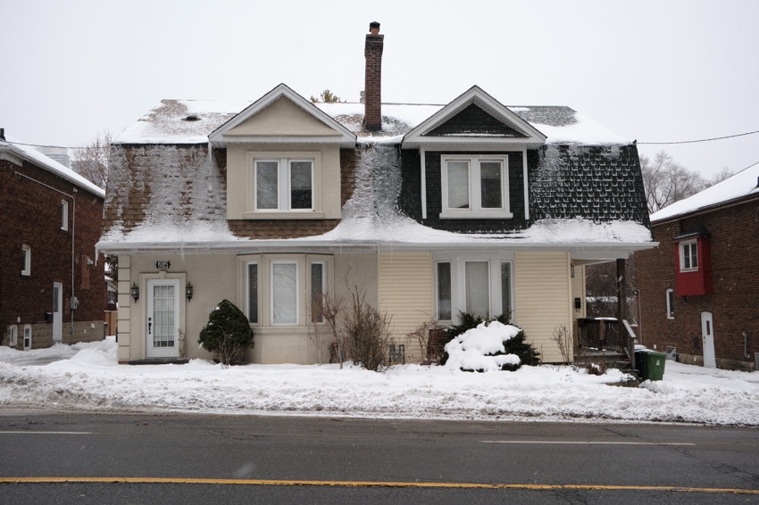 Two-story semi-detached house with twin dormers and central chimney, snow-covered roof and front yard, street in foreground.