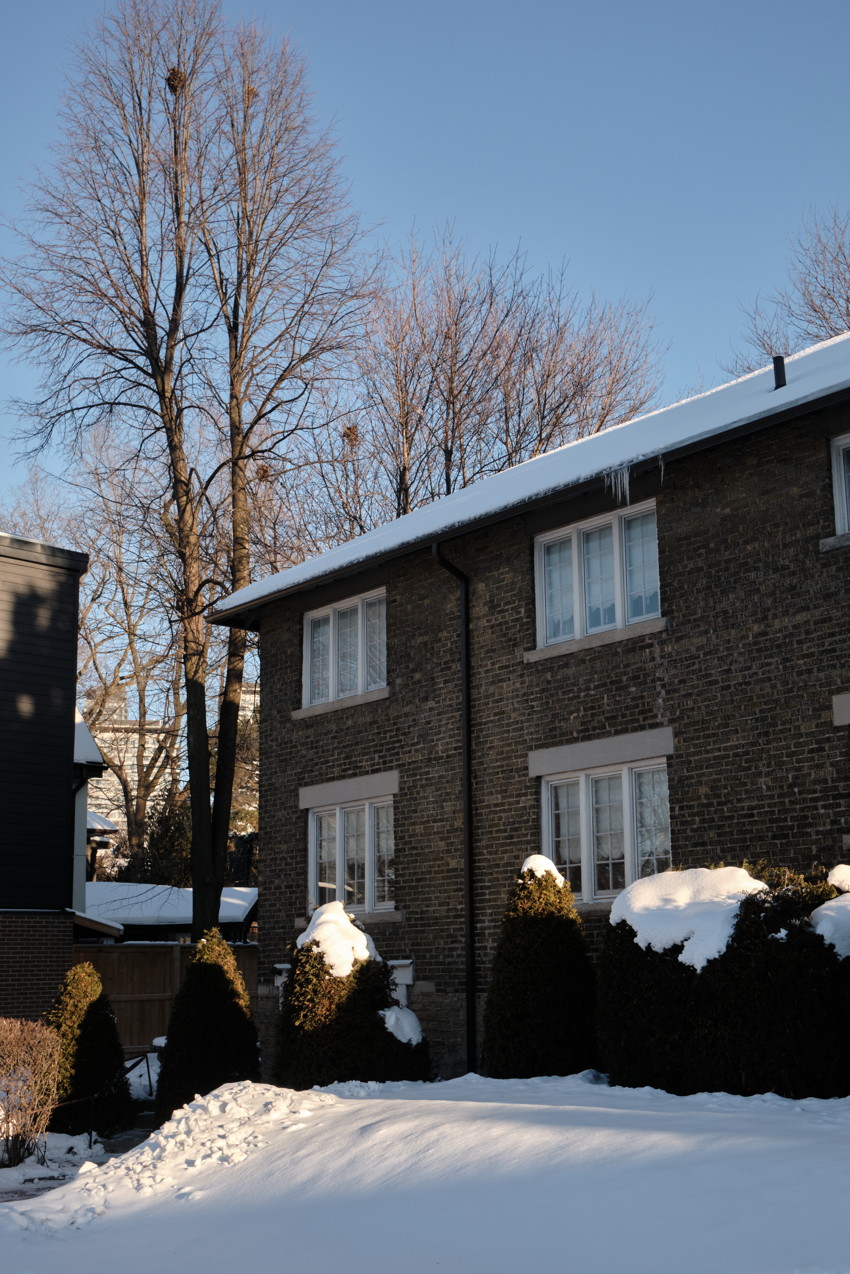 Two-story brick house with snow-covered roof and shrubs, bare trees behind, clear blue winter sky.