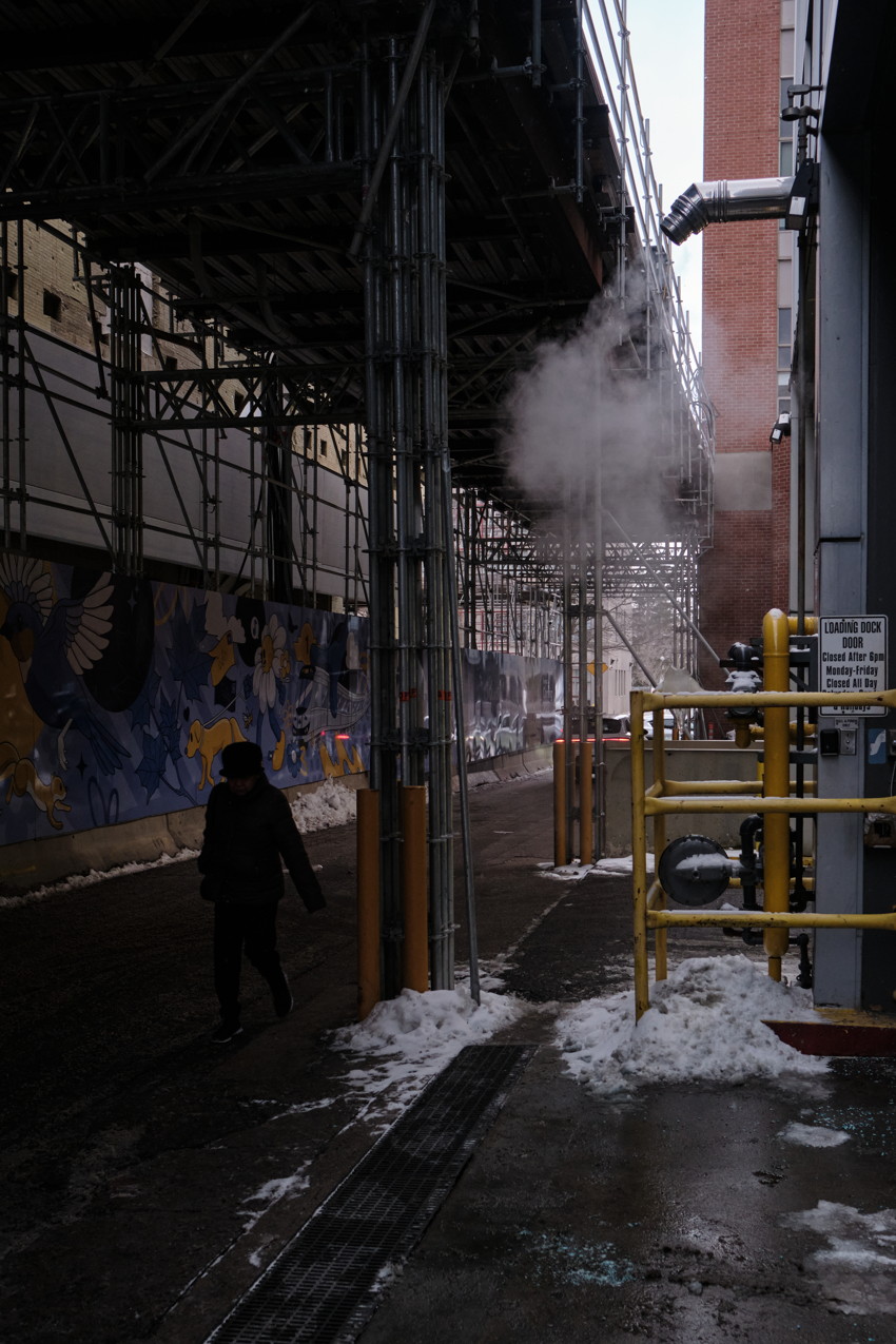Alley under scaffolding, steam plume, brick building, yellow pipes, snow, mural on left, lone walking figure.