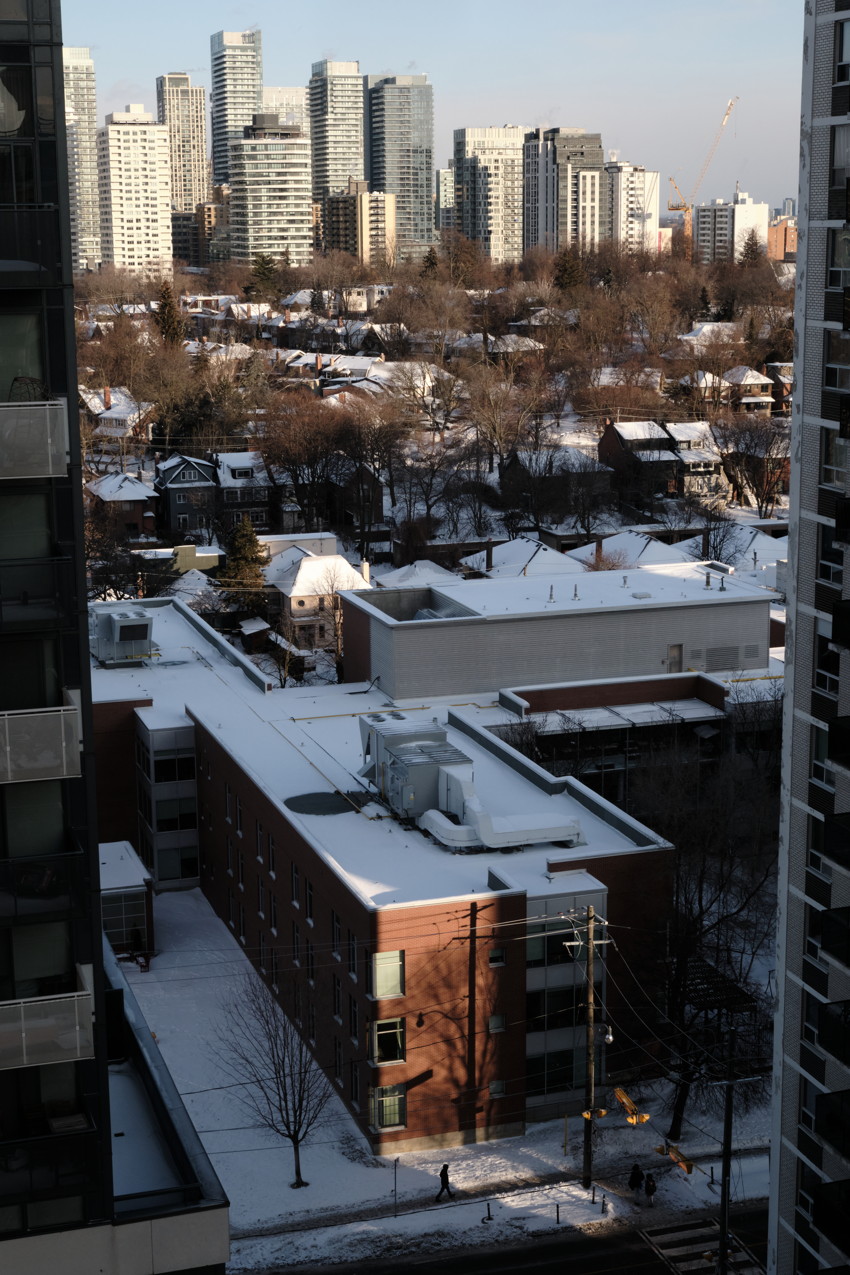 Vertical city view: snow-covered brick mid-rise in foreground, houses and high-rise towers beyond, pedestrians below.