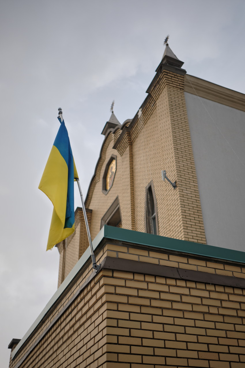 Blue-and-yellow Ukrainian flag on a pole beside a yellow-brick church facade, upward angle against cloudy sky.