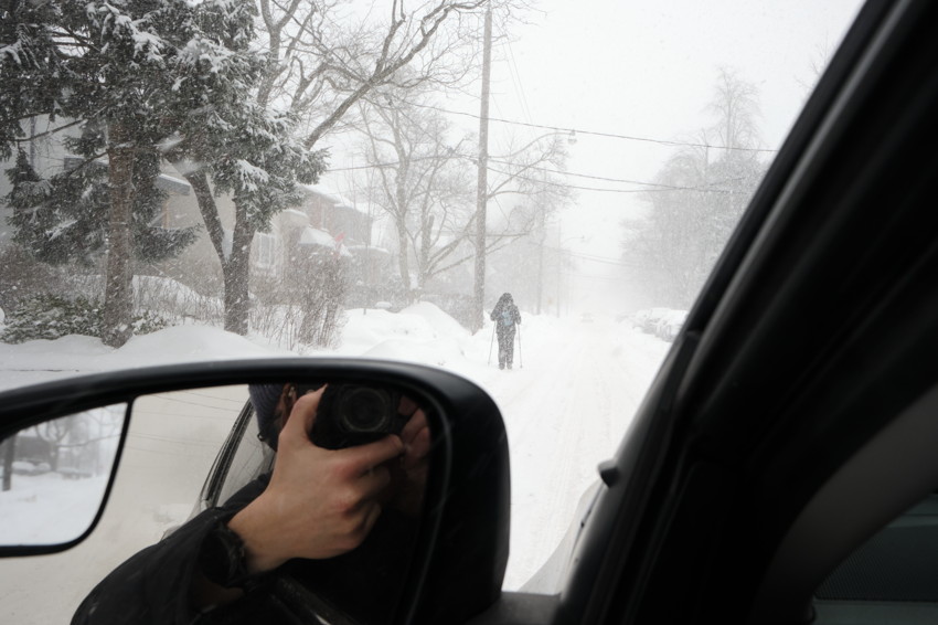 View from car in heavy snow: side mirror reflecting photographer, snowy street, lone pedestrian with walking poles.