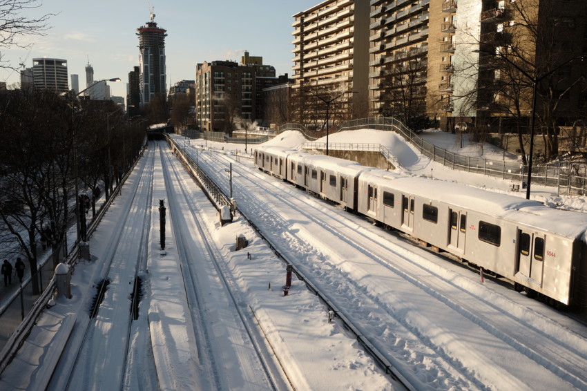 Snow-covered train tracks with parked subway train alongside high-rise apartments and distant city skyline.