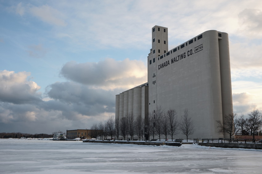 Large concrete grain elevator labeled 'Canada Malting Co.' at frozen waterfront, leafless trees and cloudy sky.