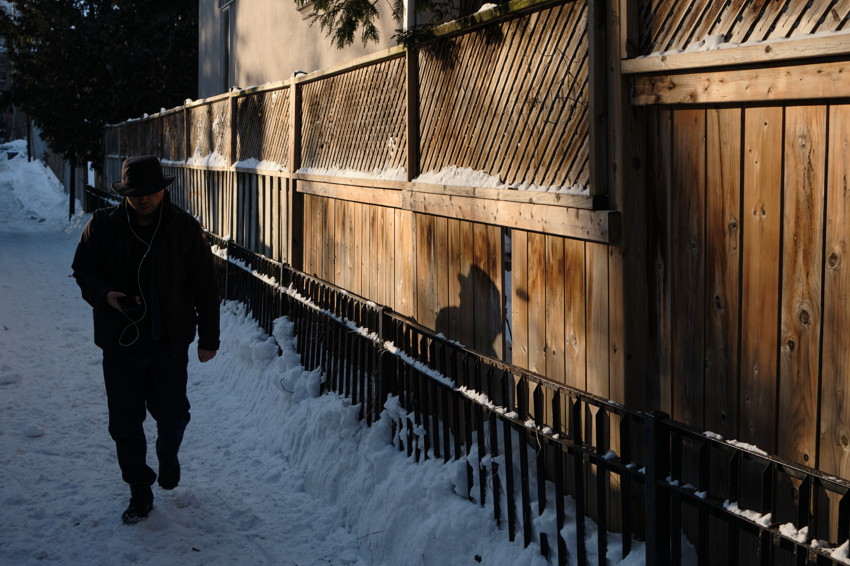 Man in hat walking along snowy sidewalk by wooden fence, holding phone; his shadow cast on sunlit fence.