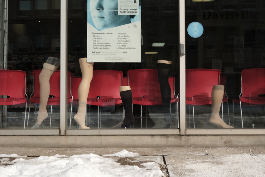 Storefront window with five mannequin lower legs wearing stockings displayed before red chairs; snowy sidewalk foreground.
