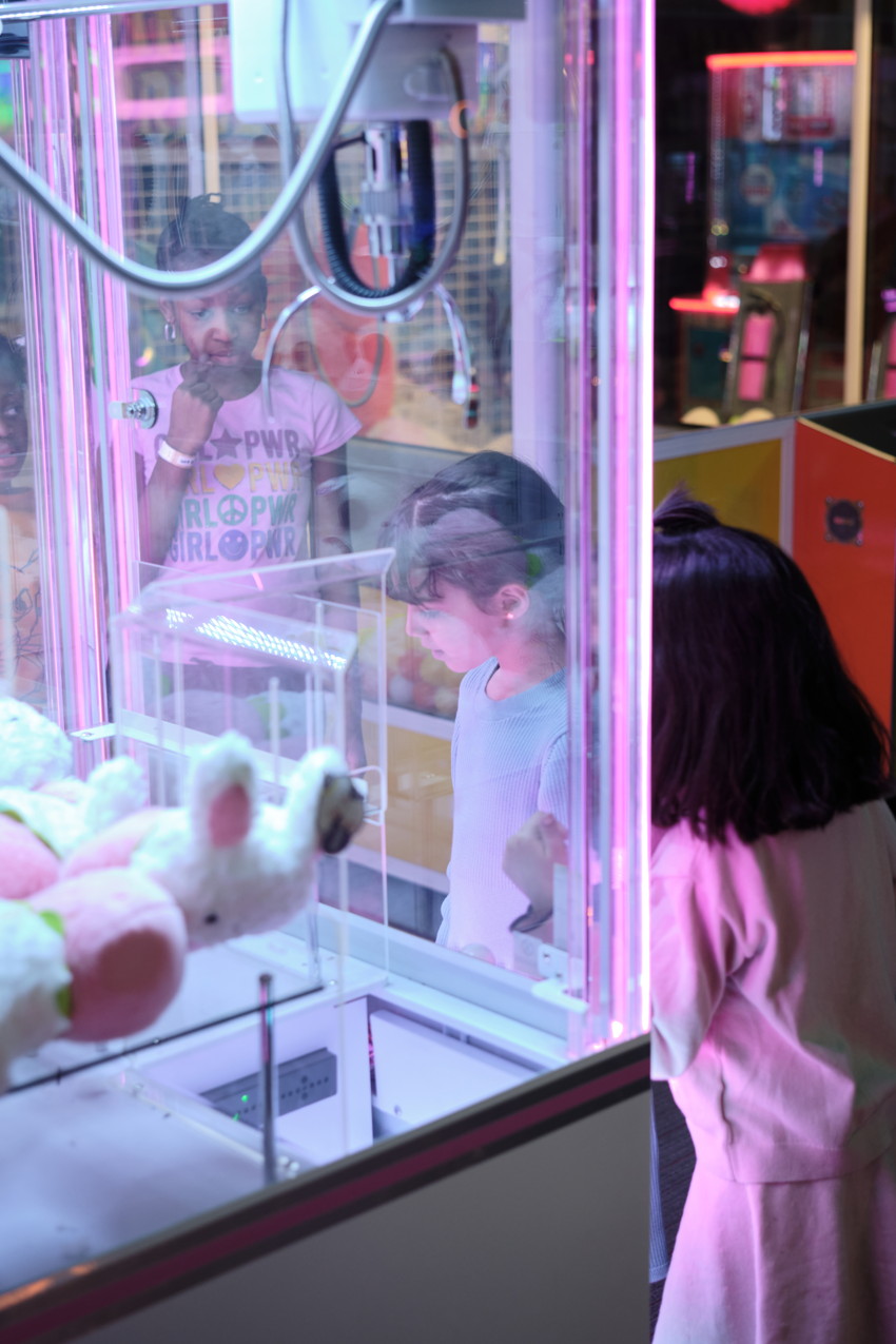 Three children watching a claw machine filled with plush toys under pink arcade lighting.