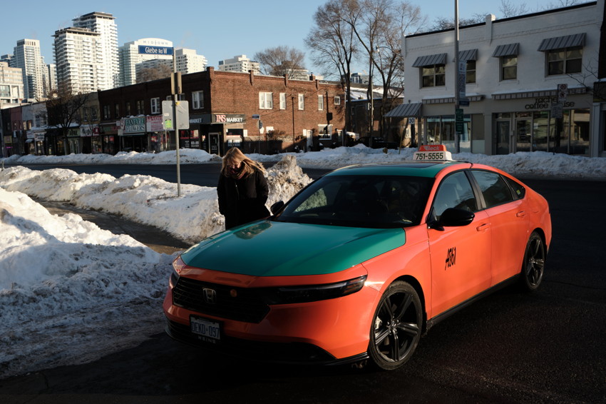 Orange-teal taxi parked on snowy street, woman nearby, storefronts and high-rise buildings in background.