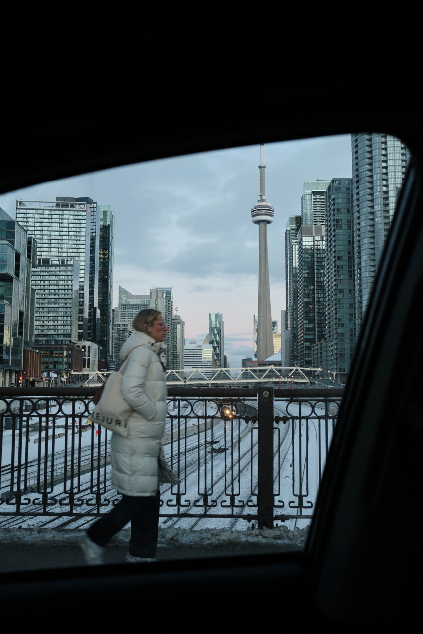 Person in white puffer coat walking past railing with CN Tower and skyscrapers, snowy tracks, framed by car window.