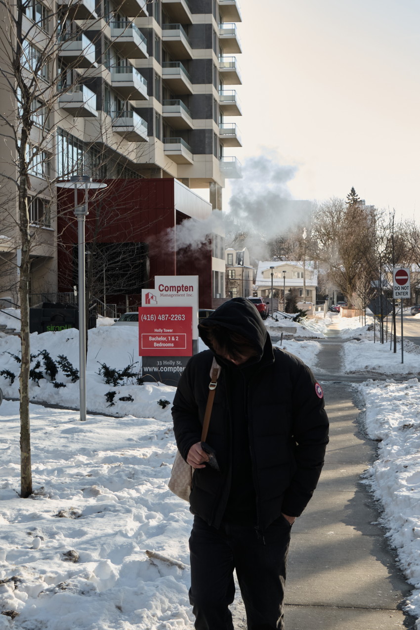Person in black hooded jacket walking on snowy sidewalk beside modern apartment building and red rental sign.
