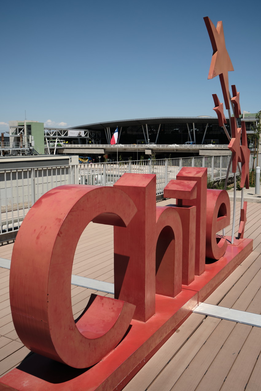 Large red 3D Chile sign on deck; metal railing, airport terminal and Chilean flag in background.