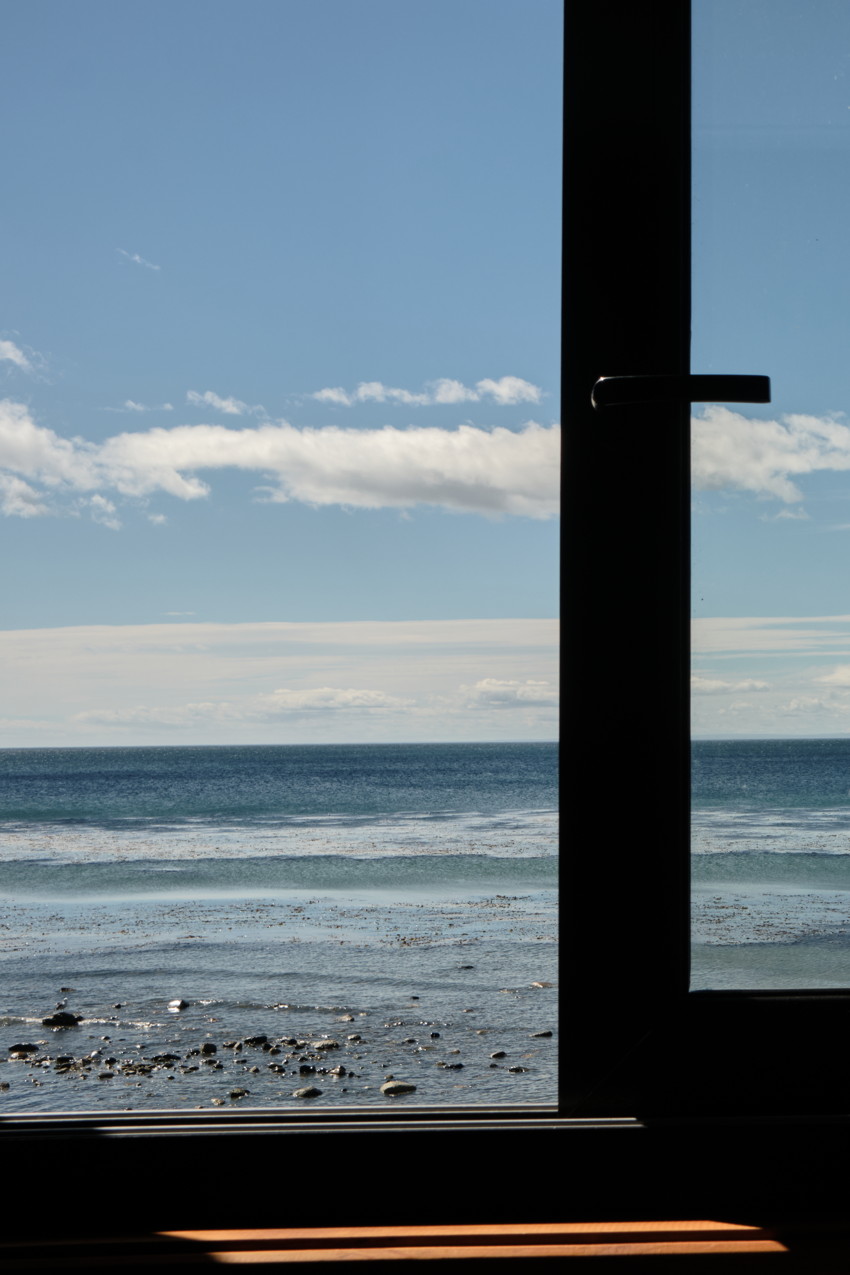 View through window: dark frame at right, rocky shoreline, calm ocean, horizon, scattered clouds, sunlight on windowsill