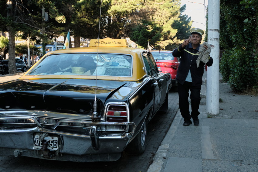 Rear three-quarter view of black-and-yellow antique taxi parked at curb; man with cap and rag standing on sidewalk.