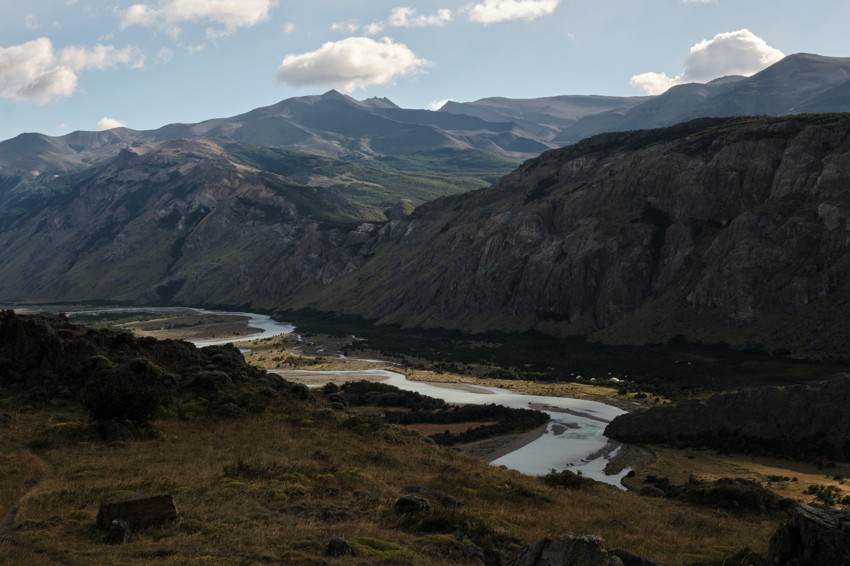 Winding river through grassy foreground, flanked by steep rocky cliffs and layered mountain ridges under clouds.