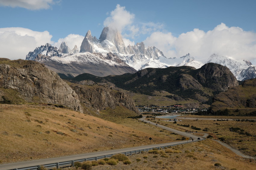 Winding road through dry grassland to a village, with rocky hills and snow‑capped jagged mountains behind.