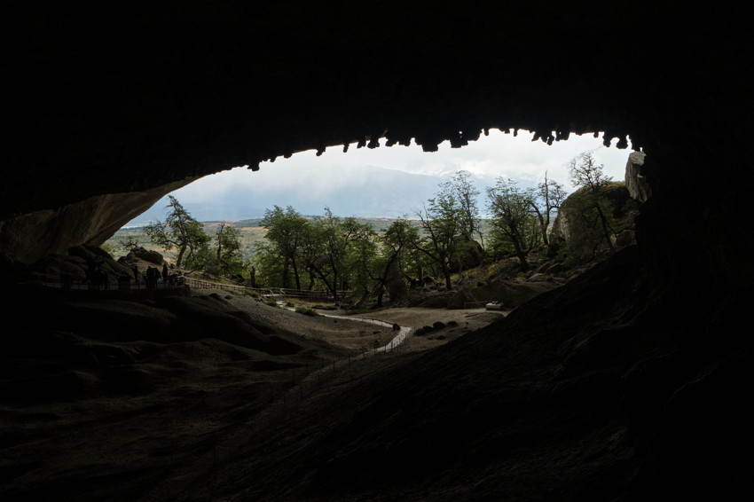 View from cave entrance framing a tree-lined valley, winding path and distant mountains under cloudy sky.