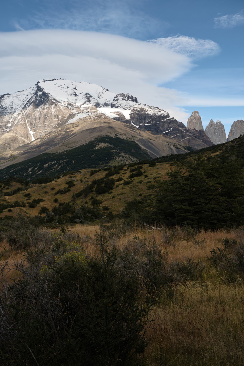 Snow-covered mountain at left, three jagged spires at right, grassy shrub foreground, lenticular cloud overhead.