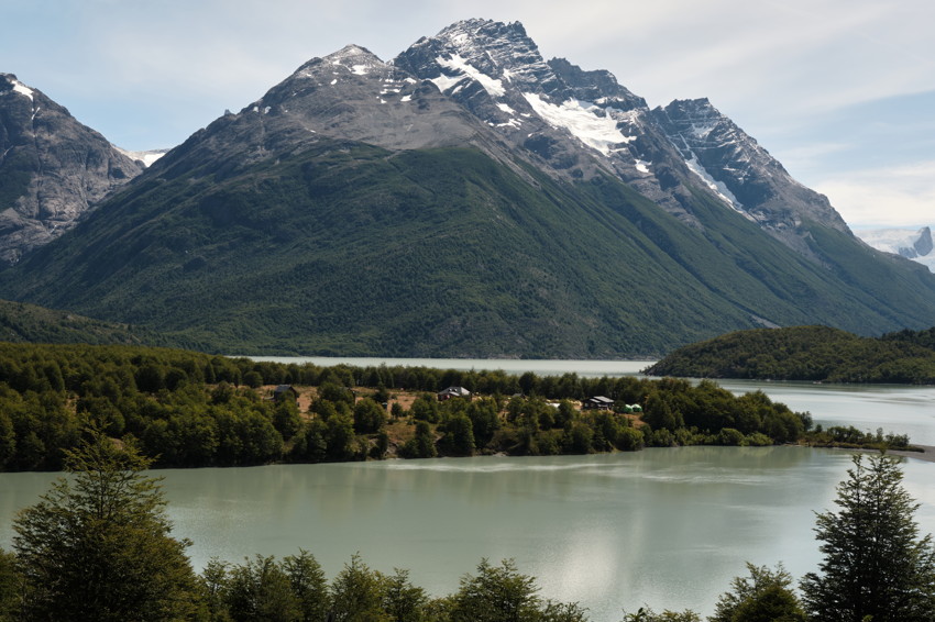 Lake with forested peninsula and scattered buildings; snow-capped mountains rise in background; foreground trees