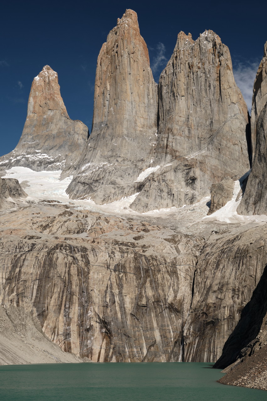 Three tall granite mountain towers with snow patches above a turquoise glacial lake, vertical rock faces.