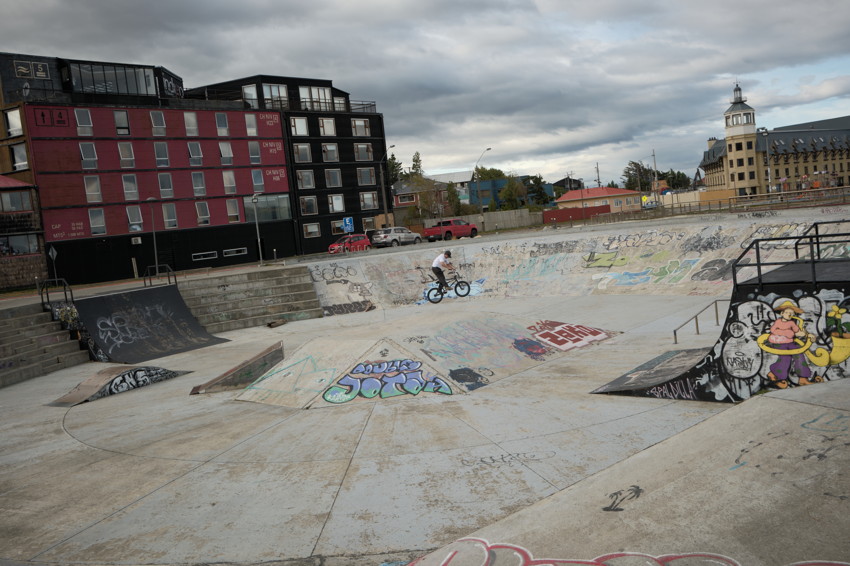 Concrete skatepark bowl with graffiti; person on BMX near center; red-and-black apartment building and cloudy sky.
