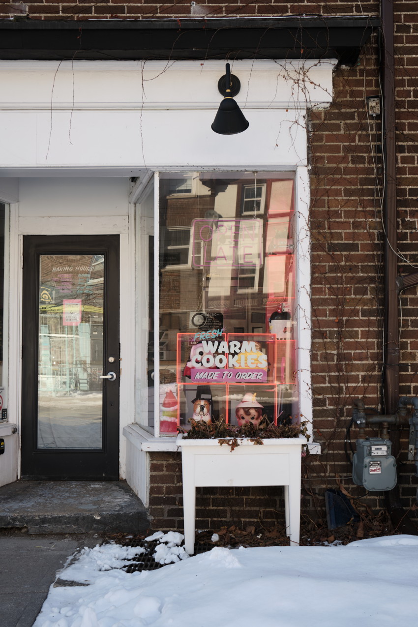 Brick storefront with glass display, black door left, neon 'Warm Cookies' sign, white planter, snow in foreground.