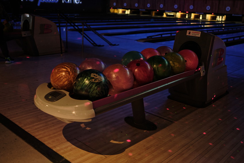 Rack of colorful bowling balls on a ball return beside lanes in a dimly lit bowling alley with wooden floor.