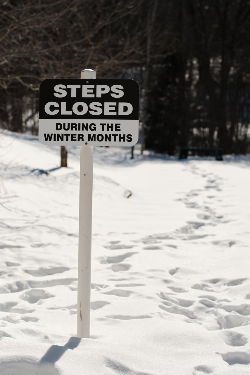 Sign on a post reading 'STEPS CLOSED DURING THE WINTER MONTHS' in snow; footprints lead toward dark treeline.
