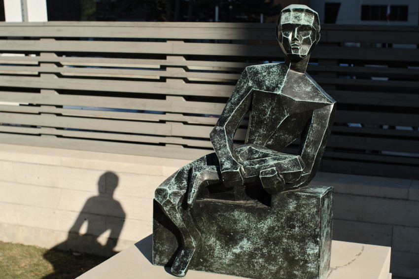 Seated geometric bronze statue with green patina on rectangular base; strong left shadow against slatted fence.