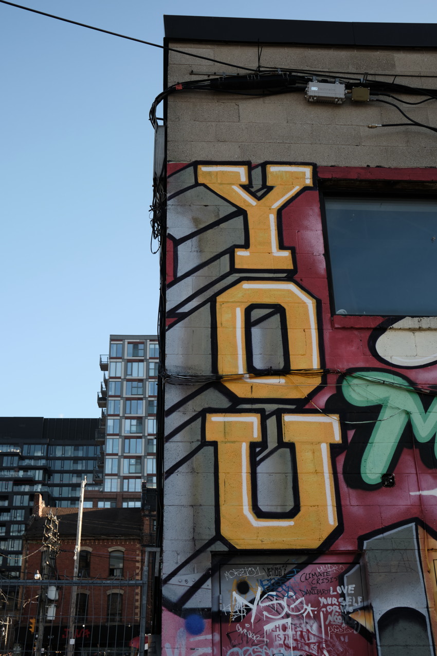 Concrete building corner with large vertical yellow graffiti spelling YOU, window, adjacent glass apartment buildings.