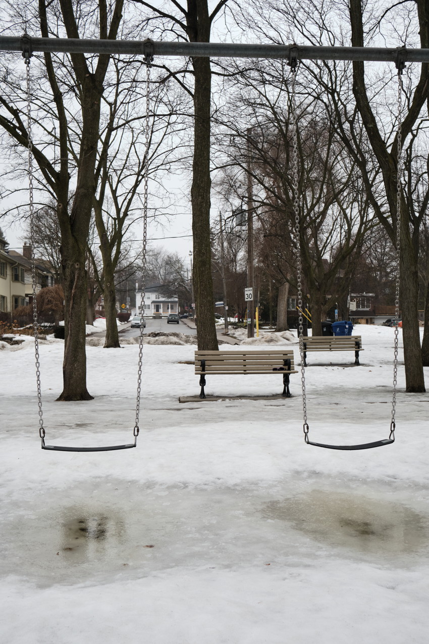Two empty swing seats in foreground over icy snow, park benches and leafless trees, houses in background.