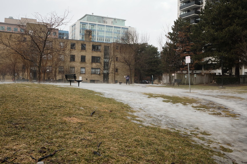 Patchy grassy foreground, icy path, empty bench left, apartment buildings background, two people walking a dog in distance.