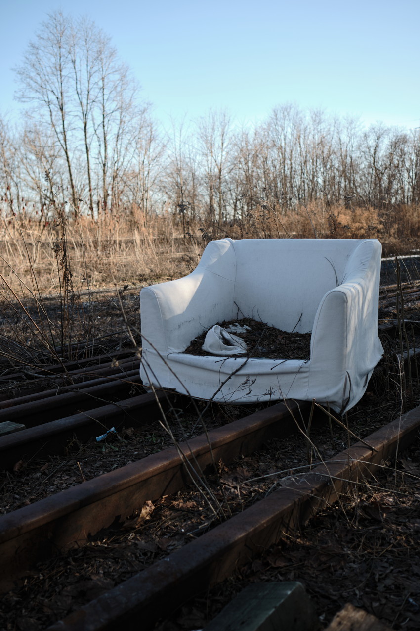 White armchair centered on rusty railway tracks in overgrown field with leafless trees and clear sky.