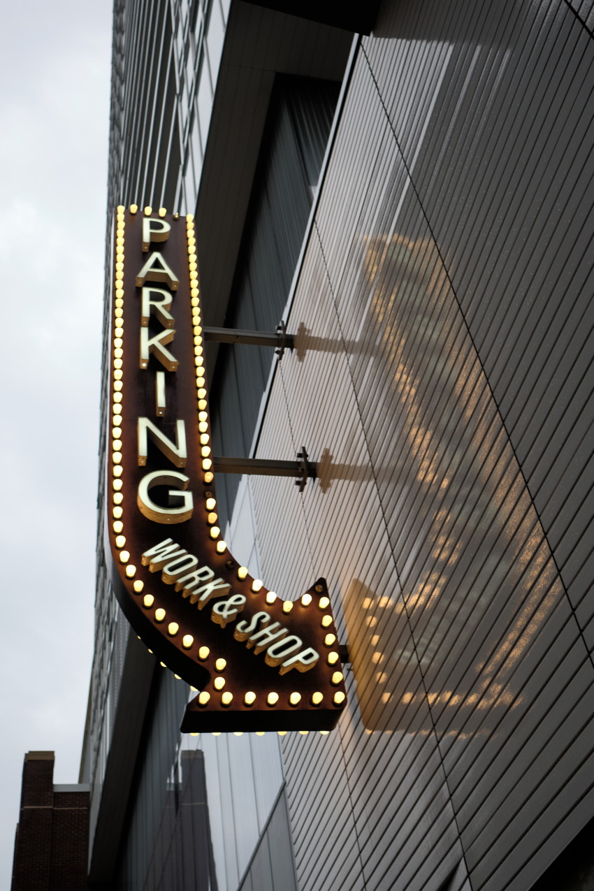 Upward view of illuminated 'PARKING' arrow sign with bulbs and 'WORK & SHOP' text, reflected on metal-clad building.