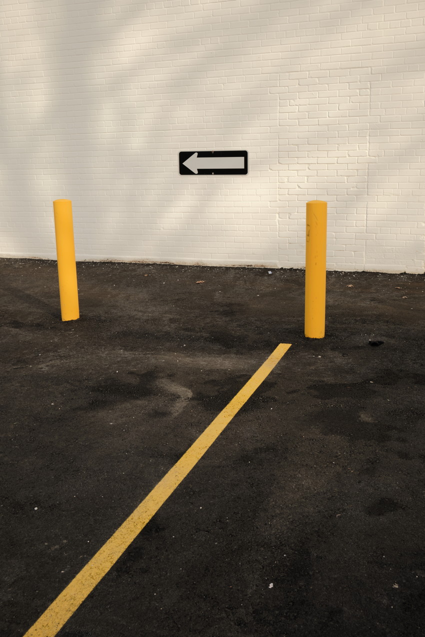 Asphalt lot with yellow stripe leading to two yellow bollards and a white brick wall with black left arrow.
