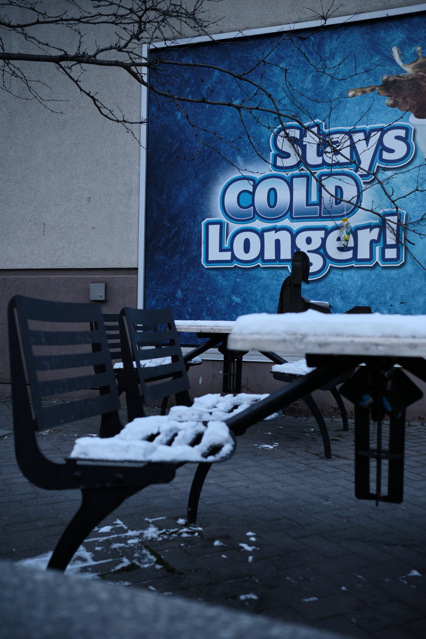 Snow-covered outdoor bench and table in foreground, blue Stays Cold Longer billboard on wall behind.