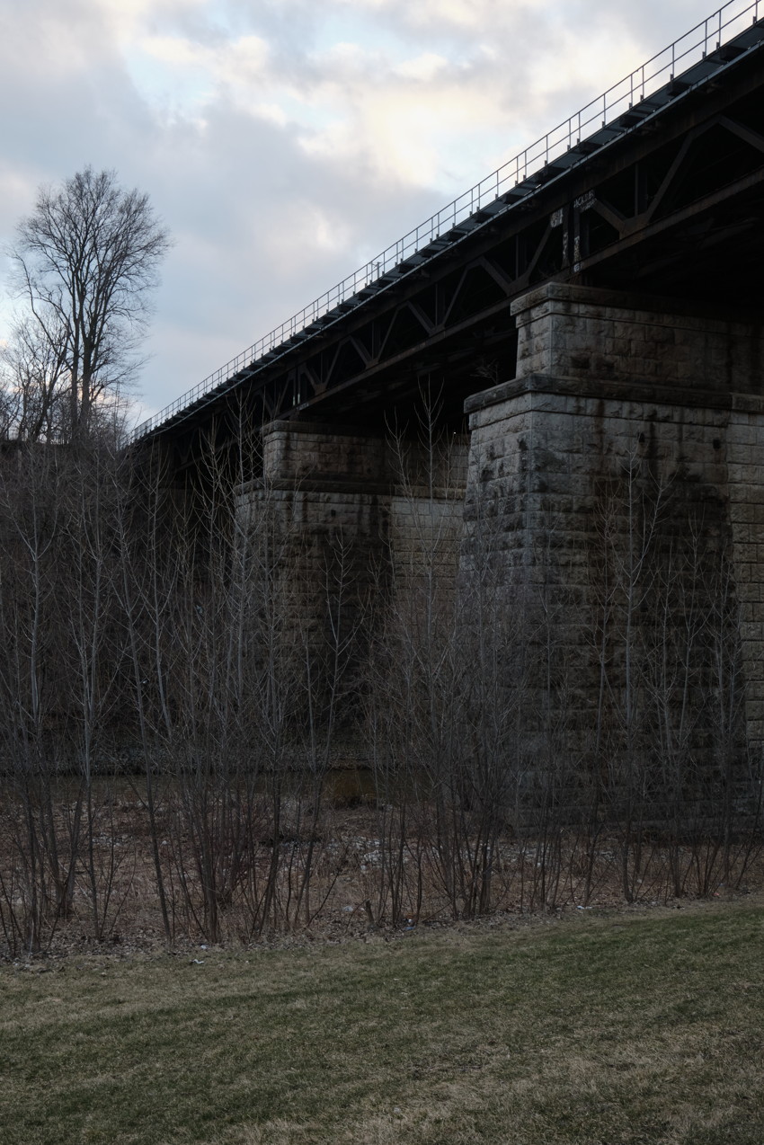 Stone railway viaduct with metal truss, four large stone piers, leafless trees and grassy foreground under cloudy sky.