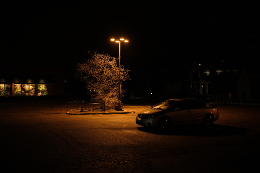 Night parking lot with lone lit streetlamp illuminating a leafless tree and bench; parked car to right.